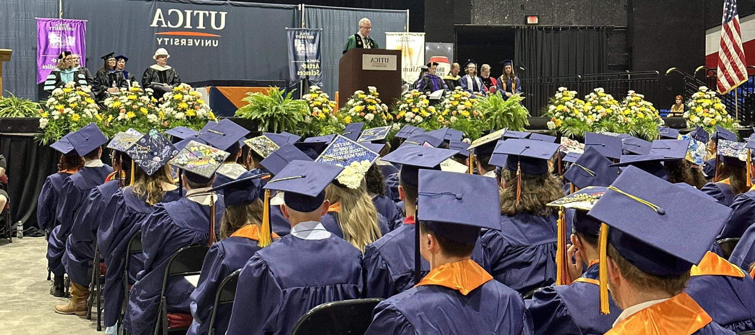 Members of the Class of 2024, in caps and gowns, sit in the Utica Memorial Auditorium as President Todd Pfannestiel speaks from a podium.